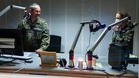 Ein Soldat und eine Soldatin stehen nebeneinander lächelnd an einem Mikrofon in einem beleuchteten Aufnahmestudio.