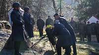 Zwei Soldaten legen auf einer Wiese an einem Gedenkstein einen Blumenkranz nieder. Um sie herum stehen Soldaten.