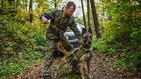 Ein Soldat steht mit einem angeleinten Hund auf einem Waldweg, im Hintergrund ein Fahrzeug
