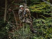Ein Soldat und ein Hund hängen zusammen an einer Seilrutsche im Wald.