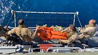 Drei Soldaten stehen an der Reling von einem Schiff und halten einen orangefarbenen Dummy in der Hand