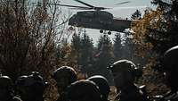 Hinter einer Gruppe Soldaten landet auf einer Waldlichtung ein großer dunkler Hubschrauber.