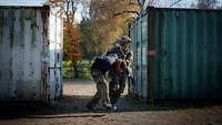 Ein Soldat rennt mit einer festgenommenen Person zwischen wie Containern über eine Sandfläche.