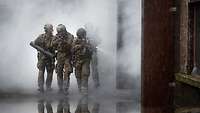 Drei Soldaten laufen mit ihrem Gewehr bei Rauch an einem Gebäude vorbei.