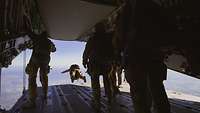 Eine Gruppe von Fallschirmspringern steht auf einer geöffneten Heckrampe. Ein Soldat ist gerade abgesprungen.