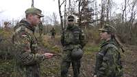 Ein Soldat und zwei Jugendliche in Uniform stehen an einem Waldweg und sprechen miteinander, dahinter laufen einige Soldaten.