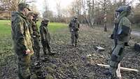 Ein Soldat erklärt einer Gruppe Jugendlicher in Uniform auf einer Waldlichtung eine militärische Ausrüstung.