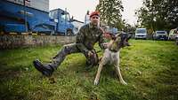 Ein Soldat kniet neben einem Hund auf einer Wiese und hält ihn fest an der Leine.