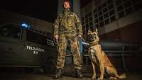 Ein Soldat steht neben einem sitzenden Hund bei Dunkelheit. Im Hintergrund steht ein Feldjäger-Fahrzeug.