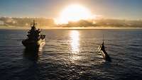 Ein Kriegsschiff und ein U-Boot fahren nebeneinander auf hoher See bei tiefstehender Sonne.