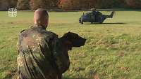 Ein Soldat hält einen Hund im Arm vor einem bereitstehenden Hubschrauber auf einer Wiese.