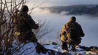 Zwei Soldaten sitzen im Schnee auf einem Berg. Sie zeichnen eine Karte und blicken ins Tal.