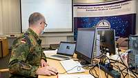 A soldier is sitting at a computer with two monitors. On one monitor an aircraft can be seen