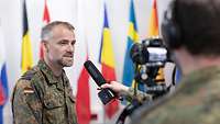 A German soldier speaks into a microphone while being filmed. Flags can be seen in the background