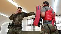 wo soldiers in hand-to-hand combat. One of them holds a mat in front of his body for protection.