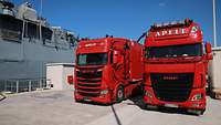 Zwei rote Lastkraftwagen stehen vor dem Schiff 
