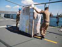 Zwei Soldaten an einer vollgepackten Palette auf dem Flugdeck eines Schiffes 