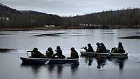 Vier jeweils Soldaten sitzen in zwei Ruderbooten auf einem See, im Hintergrund hüglige Landschaft.