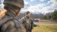 Ein Soldat mit Weste und Gewehr steht auf einer Wiese und spricht vor einer Gruppe Soldatinnen und Soldaten 