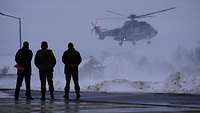 Drei Soldaten, von hinten zu sehen, stehen vor einem landenden Hubschrauber der Bundespolizei bei Schnee.