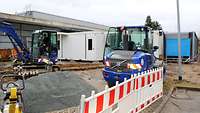 Auf einer Baustelle heben zwei kleine blaue Bagger-Fahrzeuge mehrere Gruben aus