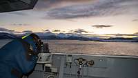 Ein Marinesoldat in blauer Arbeitsjacke schaut auf eine Bucht; im Hintergrund schneebedeckte Berge.