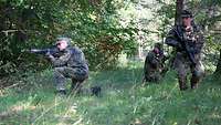 Zwei Soldaten knien im Gras mit dem Gewehr im Anschlag, einer geht mit seiner Waffe vorsichtig vorwärts.