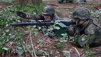 Zwei Soldaten liegen im Wald, einer hält das Maschinengewehr schussbereit im Anschlag, der andere weist ihm die Richtung zu.