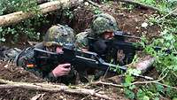 Zwei Soldaten liegen in einer Mulde im Waldboden, das Gewehr im Anschlag.