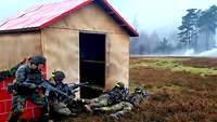 Bewaffnete Soldaten liegen und hocken im Gelände hinter einem provisorischen Haus mit rotem Dach.