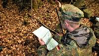 Ein Soldat kniet mit Karte und Funkgerät in der Hand auf dem Laubboden im Wald.