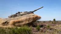 Ein Kampfpanzer vom Typ Leopard in voller Fahrt im Gelände hebt ab und wirbelt Staub auf. 
