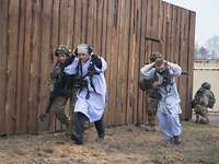 Zwei Soldaten rennen mit zwei Zivilisten vor einem Holzgebäude entlang. Ein anderer Soldat sichert shockend an der Gebäudeecke.