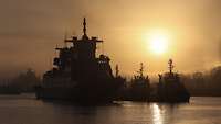 Ein Kriegsschiff und drei Schleppschiffe liegen in an der Pier im Hafen bei Nebel und tief stehender Sonne.