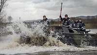 Vier Soldaten stehen auf einem Motorboot, das ein Wendemanöver auf einem Fluss macht.