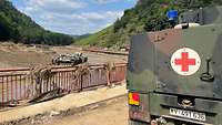 Ein Sanitätsfahrzeug der Bundeswehr steht auf einer Straße an der Ahr. Im Flussbett steht ein Bergepanzer.