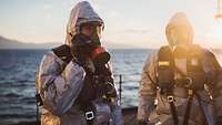 Zwei Soldaten stehen auf dem Deck eines Schiffes und tragen eine Schutzausstattung und Atemschutzmasken
