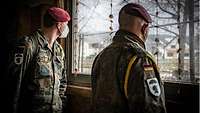 Zwei Soldaten stehen in einem mit Schlamm verschmutzten dunklen Raum und schauen aus dem Fenster. 