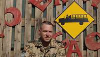 Ein Soldat steht vor einer Holzwand, an der ein gelbes rautenförmiges Schild mit schwarzem Feuerwehrauto angebracht ist