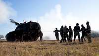 A self-propelled howitzer is standing on a meadow. Smoke is coming from the barrel; behind the vehicle, there are five soldiers.