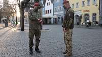 A German and a Hungarian soldier are standing on a paved town square, behind them there are rows of houses and shops.