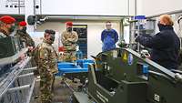 Four Hungarian soldiers are standing in a maintenance hall; two civilian mechanics in blue overalls explain their jobs.