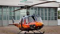 A helicopter stands in front of a large hangar.