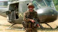 A helicopter stands on a brown meadow, in front of it is a soldier with a weapon on his chest.