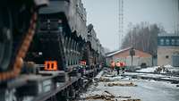 Kettenfahrzeuge stehen auf Bahnwaggons, während Bahnarbeiter in orangenen Warnwesten an diesen arbeiten