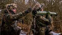 Zwei Soldaten knien im Gelände, einer zielt mit einer Panzerfaust.