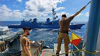 Ein Soldat schwenkt zwei Signalflaggen auf einem Kriegsschiff. Gegenüber fährt ein anderes Kriegsschiff.