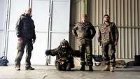 In einem Hangar stehen drei Soldaten neben einem liegenden Soldaten in Fallschirmsprungausrüstung.