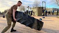 Ein Soldat hebt einen Traktorreifen an. Im Hintergrund üben Soldaten mit Sportgeräten.