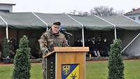 Ein Soldat in Uniform steht an einem Pult und hält eine Rede. Im Hintergrund steht ein Zelt mit Gästen.